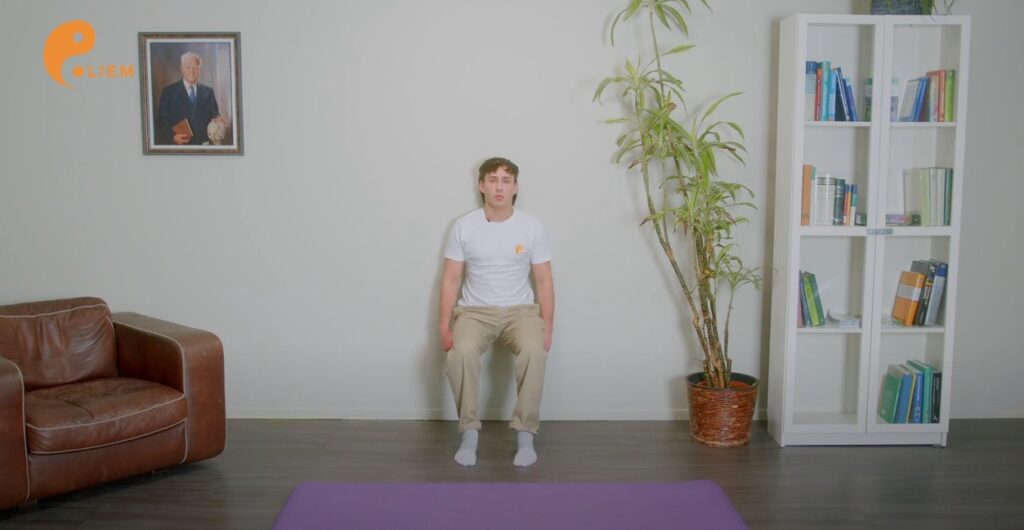 Ein Mann sitzt auf einer lila Yogamatte in einem hell erleuchteten Raum mit einem Bücherregal, einer Pflanze und einer Stuhlübung an der Wand.