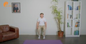 Ein Mann sitzt auf einer lila Yogamatte in einem hell erleuchteten Raum mit einem Bücherregal, einer Pflanze und einer Stuhlübung an der Wand.