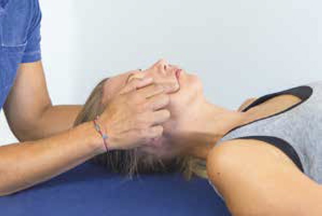 Eine Person liegt auf einem Tisch und erhält eine Gesichtsmassage. Die Hand einer anderen Person hält sanft ihr Kinn und ihren Kiefer und übt Druck auf bestimmte Punkte rund um den Modiolus aus. Die Person, die die Massage erhält, trägt ein graues Tanktop.