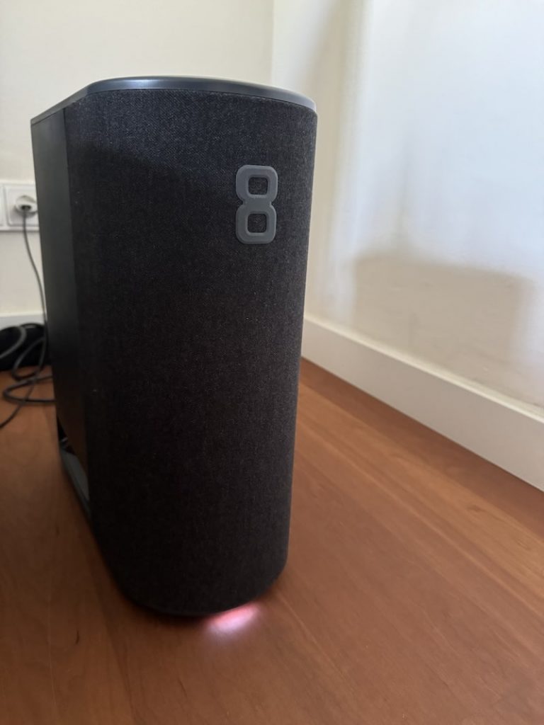 Ein dunkelgrauer, rechteckiger Smart Speaker mit der Zahl 8 auf der Vorderseite steht auf einer Holzoberfläche. Das Gerät hat eine Stoffhülle und ist an eine im Hintergrund sichtbare Steckdose angeschlossen. An der Unterseite des Lautsprechers ist ein schwaches Licht zu sehen, das ideal für einen gesunden Schlaf ist.