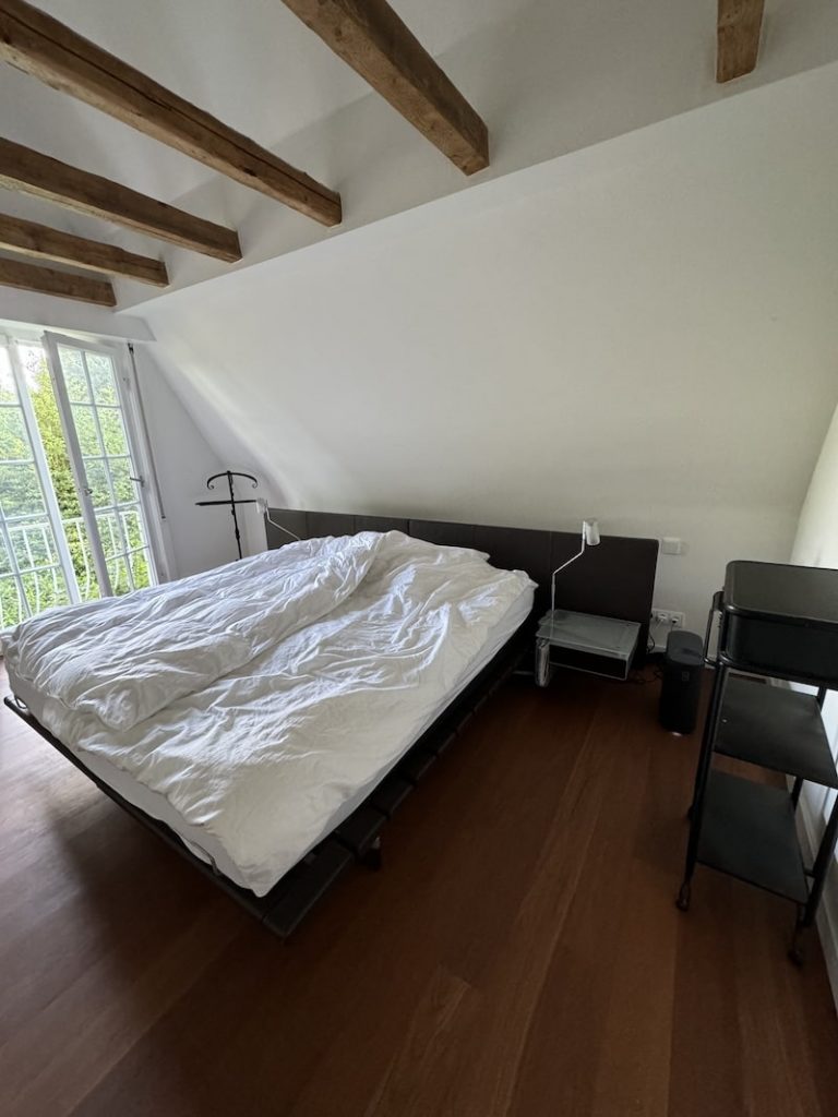 Ein Schlafzimmer mit einem großen Bett mit weißer Bettdecke, zwei Nachttischen mit Lampen, Holzboden, freiliegenden Deckenbalken, einem Kleiderständer und einem Metallregal. Das Zimmer ist mit seiner ruhigen Atmosphäre und dem natürlichen Licht, das durch das große Fenster auf der linken Seite hereinströmt, auf einen gesunden Schlaf ausgelegt.